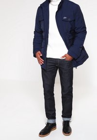 Marineblå vadderad jacka med flera fickor, ribbad vit tröja, mörk jeans, och svarta snörskor med bruna sula accenter.