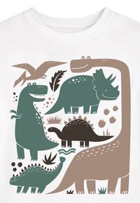 Tegneserie-dinosaurer inklusive T-Rex, triceratops, stegosaurus, pterodaktyl og brachiosaurus i grøn og brun på en hvid skjorte.