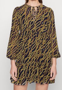 Tummansininen mekko, jossa on kultaketju-kuvio, pitkät hihat, pyöreä pääntie ja kerroksellinen röyhelöhame. Avainaukko etudetalji.