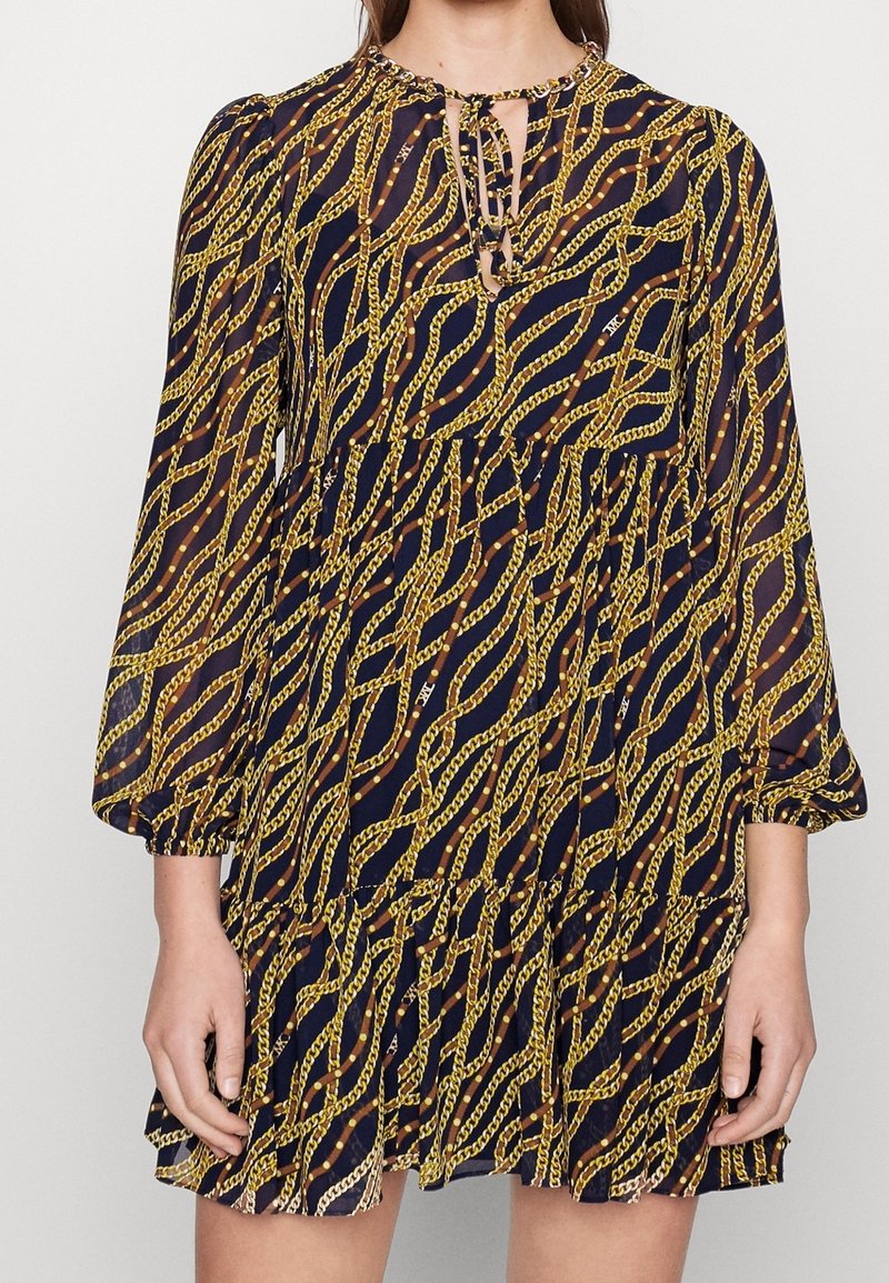 Tummansininen mekko, jossa on kultaketju-kuvio, pitkät hihat, pyöreä pääntie ja kerroksellinen röyhelöhame. Avainaukko etudetalji.