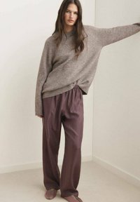 Hellbrauner, übergroßer Wollpullover mit geripptem Saum, kombiniert mit lockeren, tiefpflaumenfarbenen Hosen und hellbraunen Slippern mit grünen Akzenten.