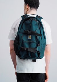 Mørke teal rygsæk med flere stropper og en frontmesh-lomme. Har et logo-patch og et slankt, holdbart stofdesign.