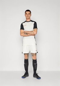 Nike-jalkapallovaruste, joka sisältää valkoisen paidan mustilla hihoilla, valkoiset shortsit, mustat sukat ja mustat kengät. Mukana logo-akit.