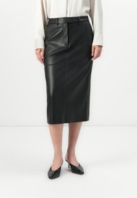 Femme portant une jupe midi crayon en cuir noir avec une taille ceinturée, un blouse blanche à manches longues et des chaussures à talons kitten noires à bout pointu.