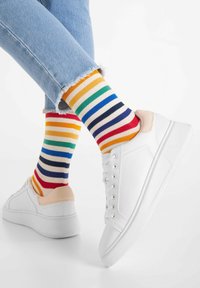 Weiße Sneakers mit einem beigen Akzent, kombiniert mit bunten gestreiften Socken in Rot, Gelb, Grün, Blau und Marineblau, zu einer Jeans.
