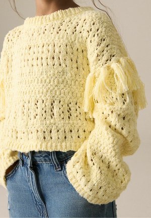 Kvinde iført en bleg gul strikket sweater med frynser på ærmerne og blå denimjeans med hænderne i lommerne.