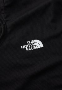 Sort stof med et rutemønster, prydet med en fremtrædende hvid logo tekst, der læser "THE NORTH FACE" og en buet, halvcirkelformet accent.