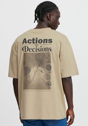 Beige oversized cotton t-shirt featuring a graphic print of text and designs on the back. Short sleeves and a relaxed fit.