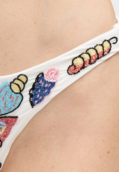 Tissu blanc avec des motifs décorés de perles colorées, comprenant un coquillage bleu et rose ainsi qu'une forme segmentée jaune et orange sur une peau nue.