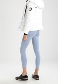 Bílá nafouknutá bunda s černými akcenty, přiléhavé světle modré džíny a černé nazouvací boty s lesklým povrchem. Bunda má kapuci.