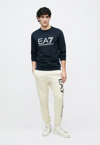 Temno modra majica s kapuco z belim logotipom "EA7 EMPORIO ARMANI", kombinirana s kremasto trenirko s črnim tiskanim logotipom. Pogled dopolnjujejo bele superge.