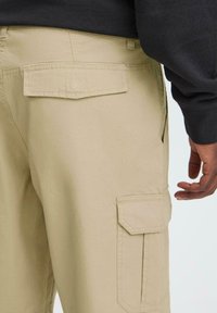 Khaki cargo bukser med flere lommer, et glat bomuldsblandingsmateriale og forstærkede syninger langs sømmene for holdbarhed.