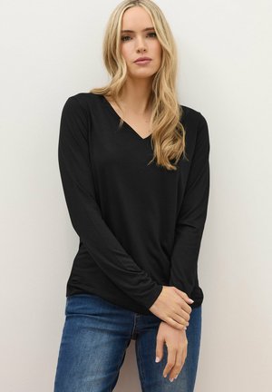 Blonde Frau, die ein schwarzes, langärmliges V-Ausschnitt-Oberteil und blaue Jeans trägt, steht vor einer schlichten, hellen Wand.