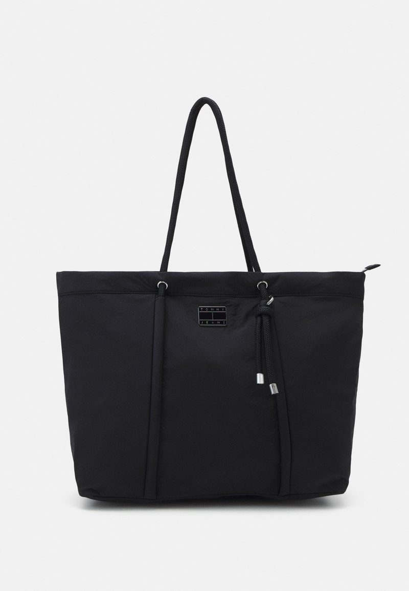Black Tommy Jeans tote bag with long rope handles, front logo plaque, and zipper closure on white background.
