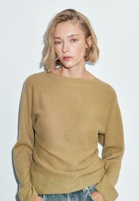 Frau mit kurzen blonden Haaren, die einen beigen Pullover und blaue Jeans trägt, steht vor einem schlichten weißen Hintergrund mit den Händen in den Taschen.