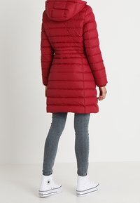 Röd puffjacka med huva, med horisontell sömnad och en längre design. I kombination med grå skinny jeans och vita högskurna sneakers.