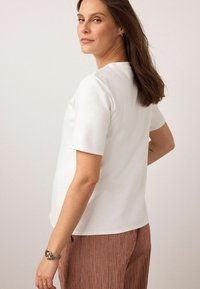 Weißes Kurzarmoberteil mit klassischem Schnitt, glatter Textur und rundem Ausschnitt, kombiniert mit karierten braunen Hosen.