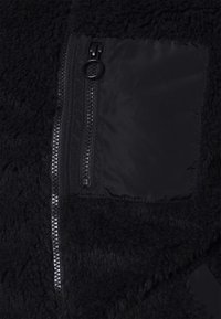 Svart fleecetröja med en mjuk, texturerad utsida och ett jämnt tygfack med dragkedjestängning. Detaljerna inkluderar metalliska dragkedjor.