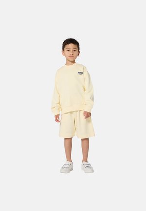 Sweat-shirt jaune en coton et short assorti. Le sweat-shirt présente un logo sur la poitrine, une coupe décontractée et un col côtelé. Le mannequin porte des chaussures blanches.
