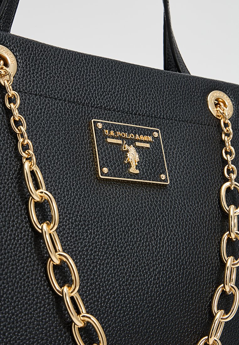 Sac en cuir noir texturé avec des accents en chaîne dorée et une plaque logo rectangulaire avec "U.S. Polo Assn." embossé.