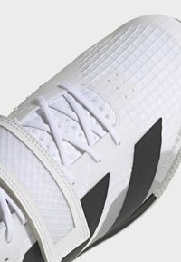 Bijela sportska cipela s teksturiranim gornjim dijelom, uzorkom mreže, tankim vezicama i crnim akcentom sa strane. Ima čičak traku za dodatnu potporu.