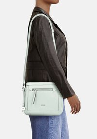 Borsa crossbody in pelle verde menta chiaro con tasca frontale con zip, tracolla regolabile e logo impresso sul davanti. Texture liscia.