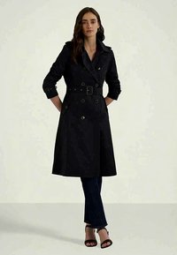 Schwarzer Trenchcoat mit doppelt geknöpftem Design, breitem Kragen, tailliertem Gürtel und langen Ärmeln; kombiniert mit dunklen Jeans und Absatzsandalen.