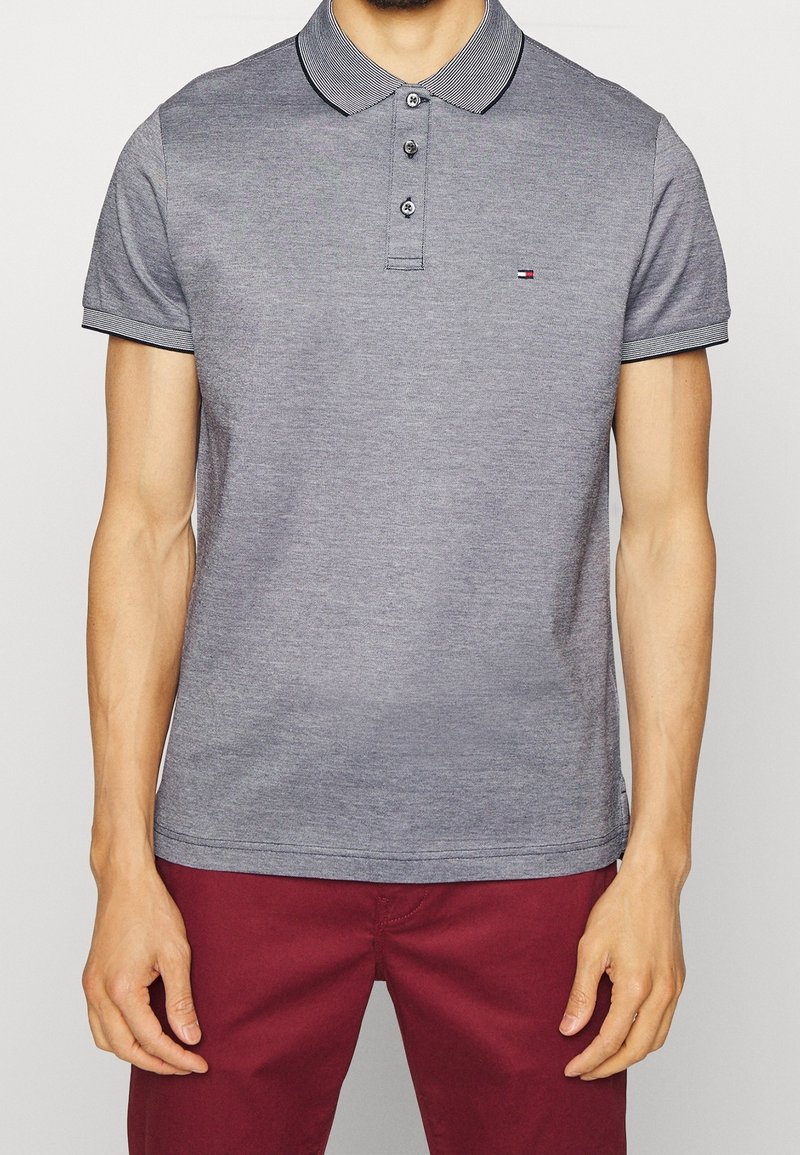 Šedá polo tričko s pruhovaným límcem a trojicí knoflíků; krátké rukávy s pruhovanými akcenty; nošeno s burgunskými kalhotami.