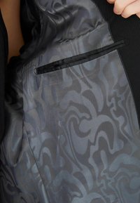 Blazer negru cu o căptușeală netedă, cu un model gri, având spirale. Include un buzunar negru cu bordură pe partea interioară stângă.