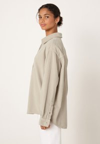 Mladá žena s kudrnatými vlasy nosí oversize béžovou kordurovou košili a bílé kalhoty, stojí v profilu na jednoduchém světlém pozadí.