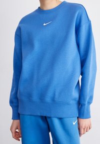 Person med en blå Nike-tröja och matchande blå Nike-byxor, som visar överkroppen och del av armarna mot en enfärgad bakgrund.