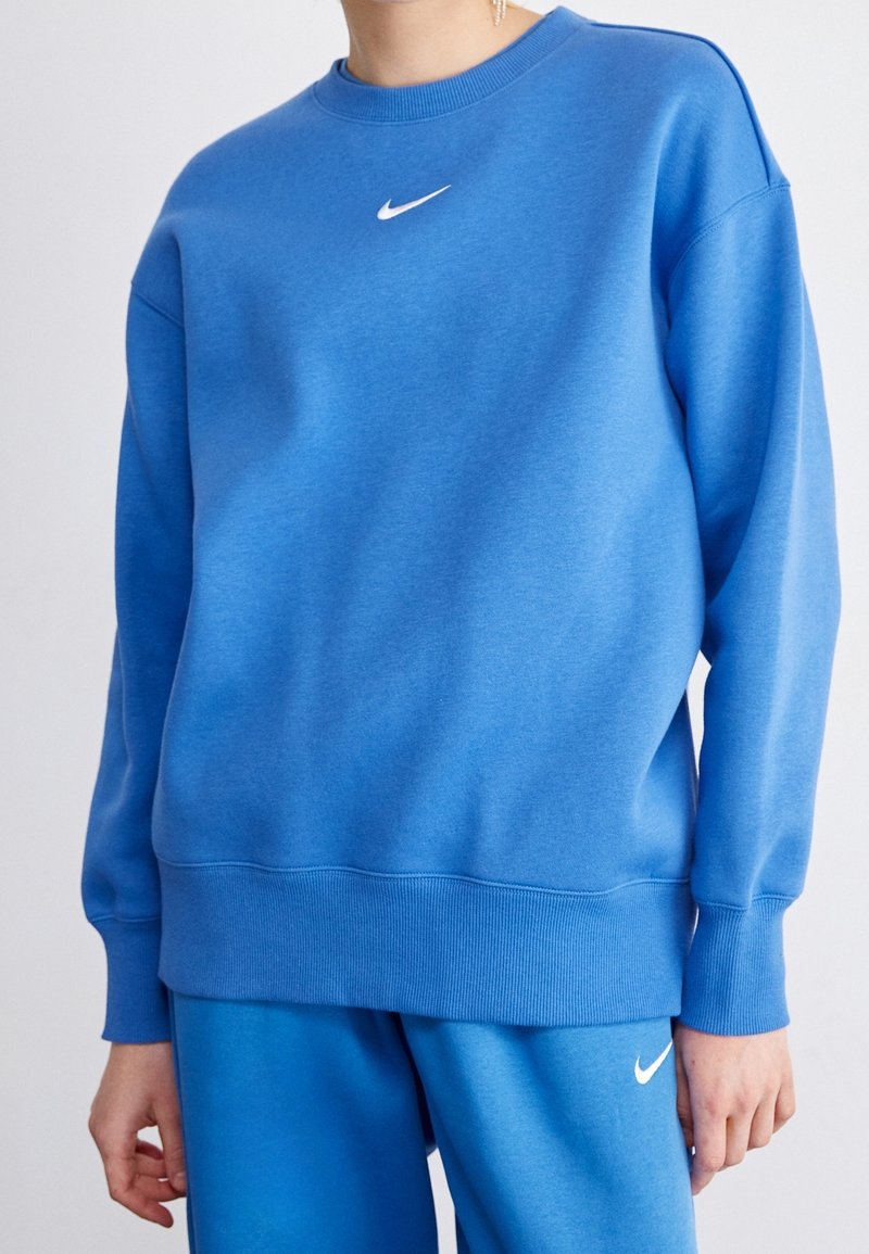 Person med en blå Nike-tröja och matchande blå Nike-byxor, som visar överkroppen och del av armarna mot en enfärgad bakgrund.