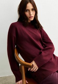 Burgunderfärgad tröja med lös passform, långa ärmar och ribbade muddar, tillverkad av mjukt material, i sällskap med en trästol.