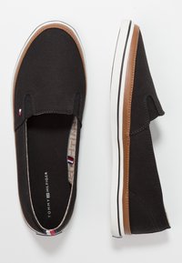 Černé plátěné slip-on boty s hnědým syntetickým koženým detailem. Bílá pryžová podrážka má černý pruh. Vnitřek je ozdoben vzorovanou podšívkou.