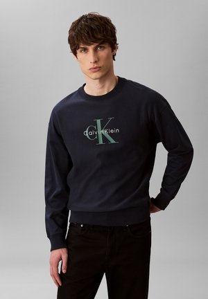 Giovane uomo con capelli castani che indossa una felpa blu navy Calvin Klein con grande logo "ck" e pantaloni neri, in piedi davanti a uno sfondo grigio.