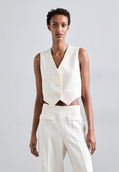 Débardeur blanc court avec un décolleté en V, comportant des perforations décoratives, trois boutons, et associé à un pantalon taille haute assorti.