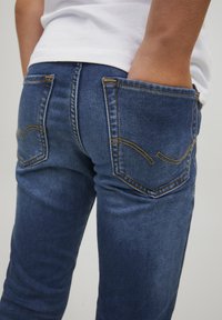 Tummansinisiä farkkuja, joissa on sukkamainen muotoilu. Niissä on kaksi takataskua, joissa on keltaista ompelua ja hienovaraisia kaarevia kuvioita.