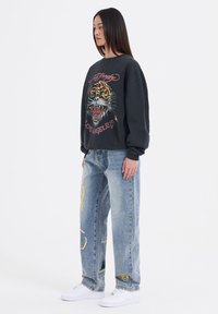 Zwarte sweatshirt met tijgerafbeelding en "Los Angeles" tekst, gecombineerd met lichtblauwe losse jeans met gele en groene ontwerpen, en witte sneakers.