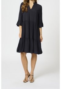 Dunkelblaues Kleid mit drei Viertelärmeln und Rüschendetails sowie einem V-Ausschnitt. Mehrlagiges Design, das bis zu den Knien reicht. Getragen mit beigen Riemchensandalen.