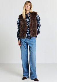 Braune Kunstpelzweste über einer blau gemusterten Bluse mit Rüschenärmeln. Kombiniert mit hellblauen Jeans mit weitem Bein und dunkelbraunen spitzen Schuhen.