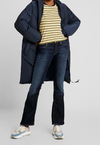 Marine blauwe pufferjas over een geel en zwart gestreept shirt, gecombineerd met donkere wijde spijkerbroeken en multicolore sneakers.