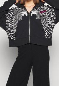 Hoodie noir court avec zip, présentant des motifs graphiques en blanc et rose. Tissu doux avec une coupe décontractée et une poche kangourou.
