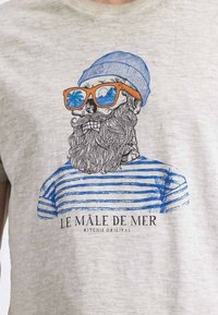 T-shirt gris avec un graphisme représentant un crâne avec une barbe portant des lunettes de soleil et un bonnet bleu, orné de rayures bleues et de texte en dessous.