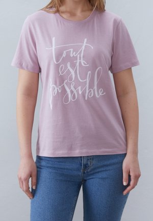 Kobieta ubrana w jasnofioletowy t-shirt z białym napisem „tout est possible” w stylu pisma odręcznego, w połączeniu z niebieskimi dżinsami, na jednolitym jasnym tle.