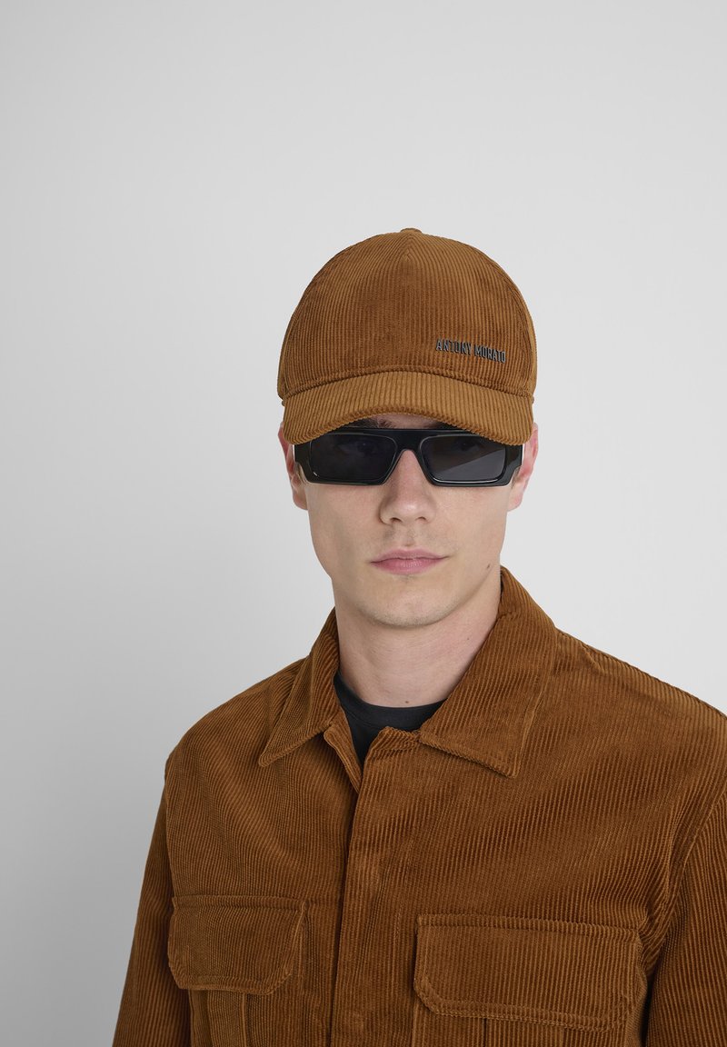 Hombre con chaqueta de pana marrón y gorra a juego, con gafas de sol negras rectangulares frente a un fondo gris.