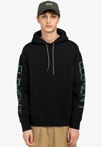 Sudadera negra con un bolsillo frontal, cordones blancos y verdes, y diseños verdes impresos a lo largo de la manga izquierda. El modelo lleva pantalones beiges.