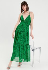 Groene maxi-jurk met een diepe V-hals, dunne banden en gelaagde stroken met een bladerenpatroon. Model draagt zilveren sandalen en staat naast een doorzichtige stoel.