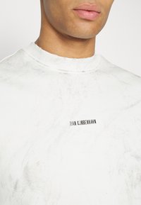 Bílá mikina s mramorovanou texturou, žebrovaným výstřihem a černým vyšitým logem značky "HAN KJABENHAWN" na přední straně.