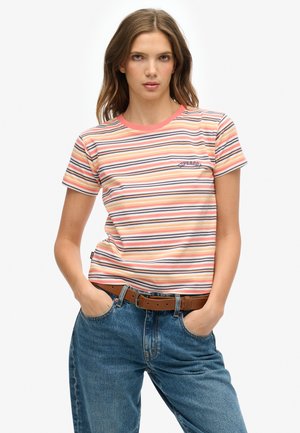 Mujer con camiseta de manga corta a rayas en tonos melocotón, blanco y azul marino, vaqueros azules y cinturón marrón, de pie con las manos en los bolsillos.