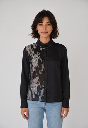 Donna con capelli corti e scuri che indossa una camicia nera con bottoni e motivo astratto grigio da un lato e jeans azzurri, in piedi davanti a uno sfondo grigio.